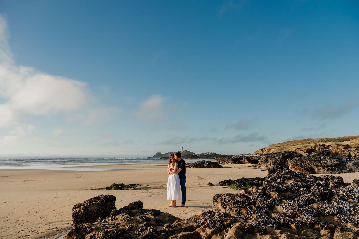 Couple embracing on Godrevy beach with rocky shores and blue skies in a romantic coastal setting.