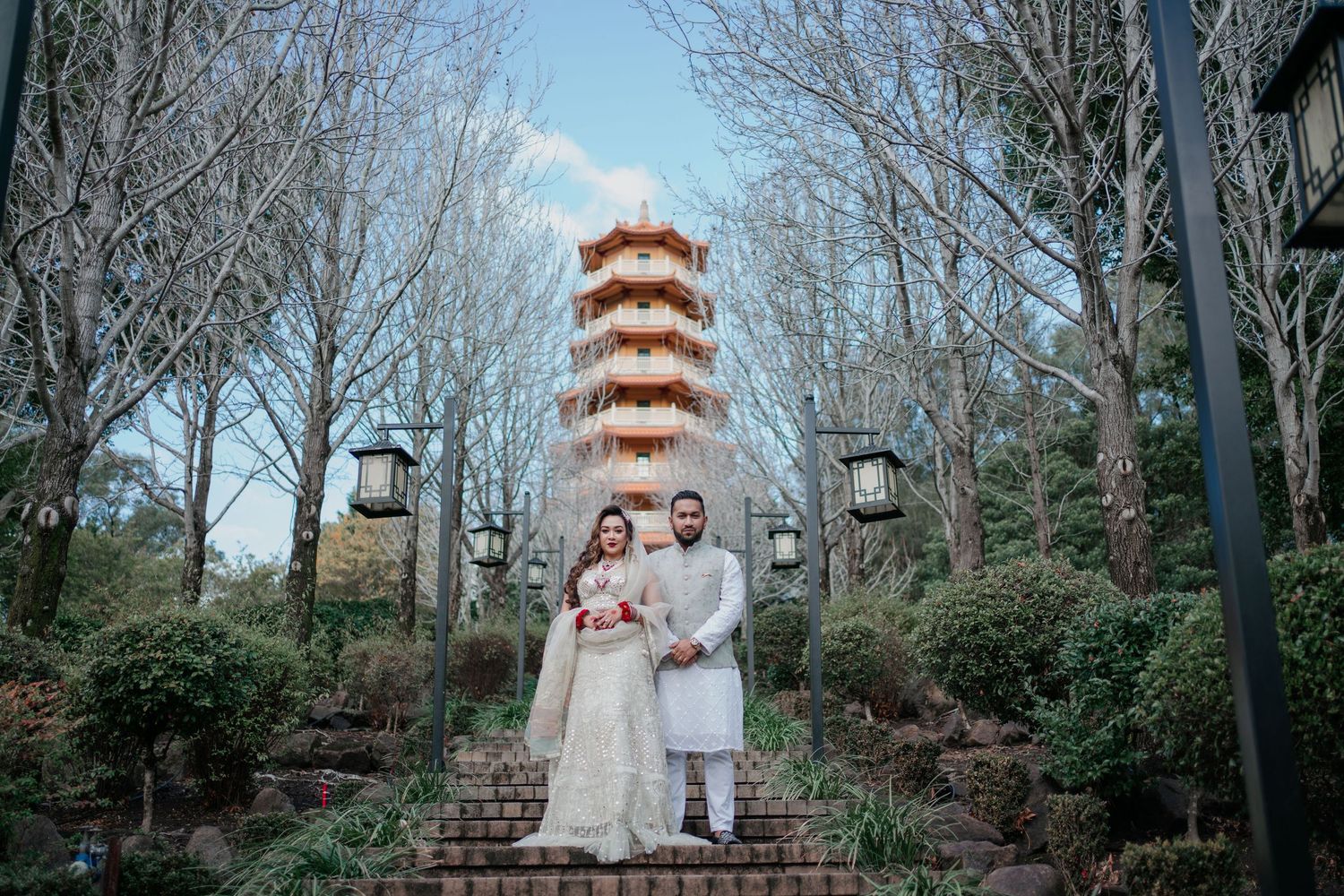 Couple in traditional wedding attire poses on stone steps in a garden with a tiered pagoda tower and bare winter trees behind them.