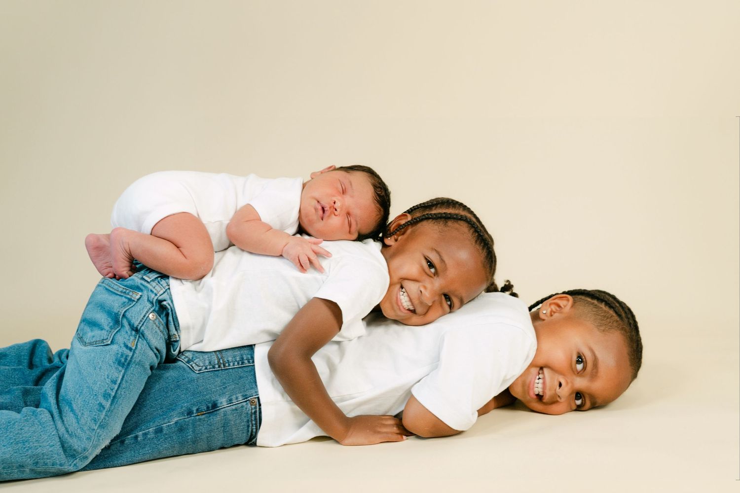 Three children pose together, two older kids in white shirts and jeans lying down with a smiling newborn baby on top.
