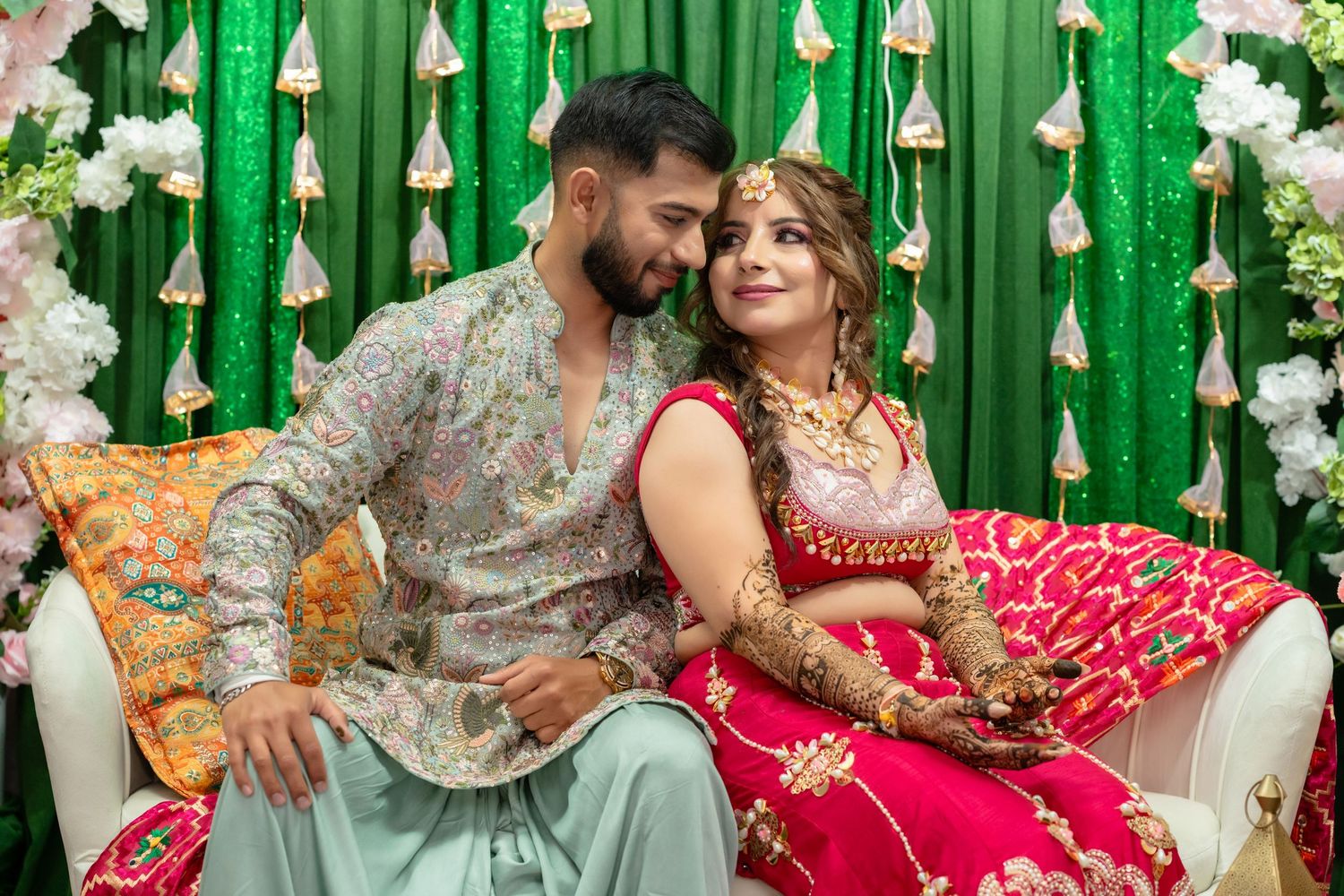 Couple in traditional South Asian wedding attire, bride in red lehenga and groom in embroidered kurta, seated against green floral backdrop.