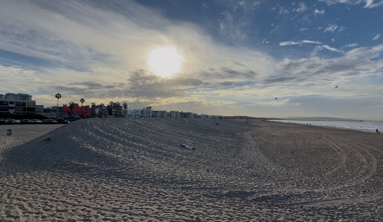 Sun shining through clouds over a wide sandy beach with tire tracks and colorful buildings and palm trees along the distant shoreline.