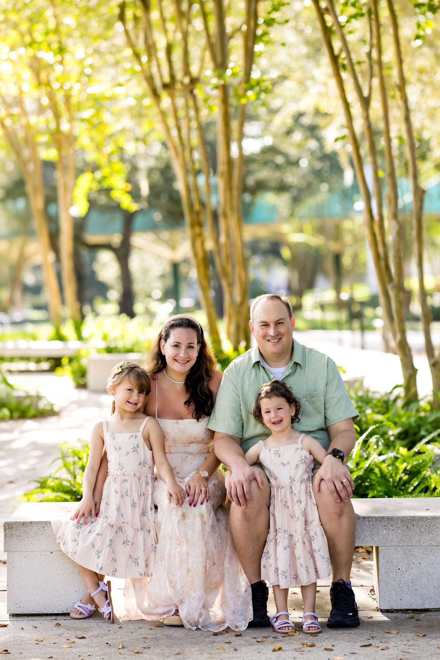 Family of four smiling together on a bench in a sunny park, two girls in matching floral dresses with parents.