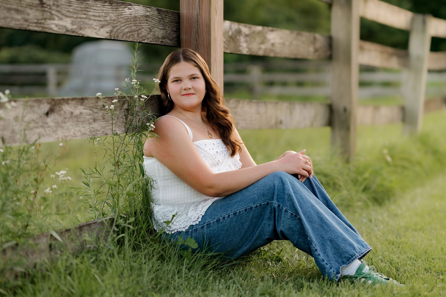 A young woman in a white top and jeans sits smiling against a wooden fence in a lush green field.