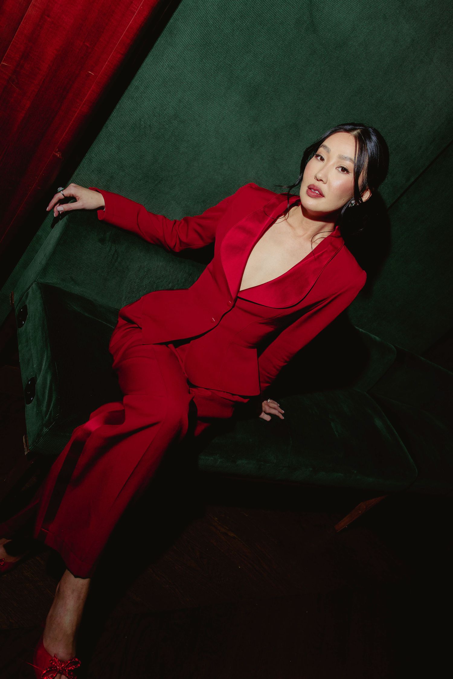 Bride in red pantsuit posing elegantly on green velvet chair against dark green wall.