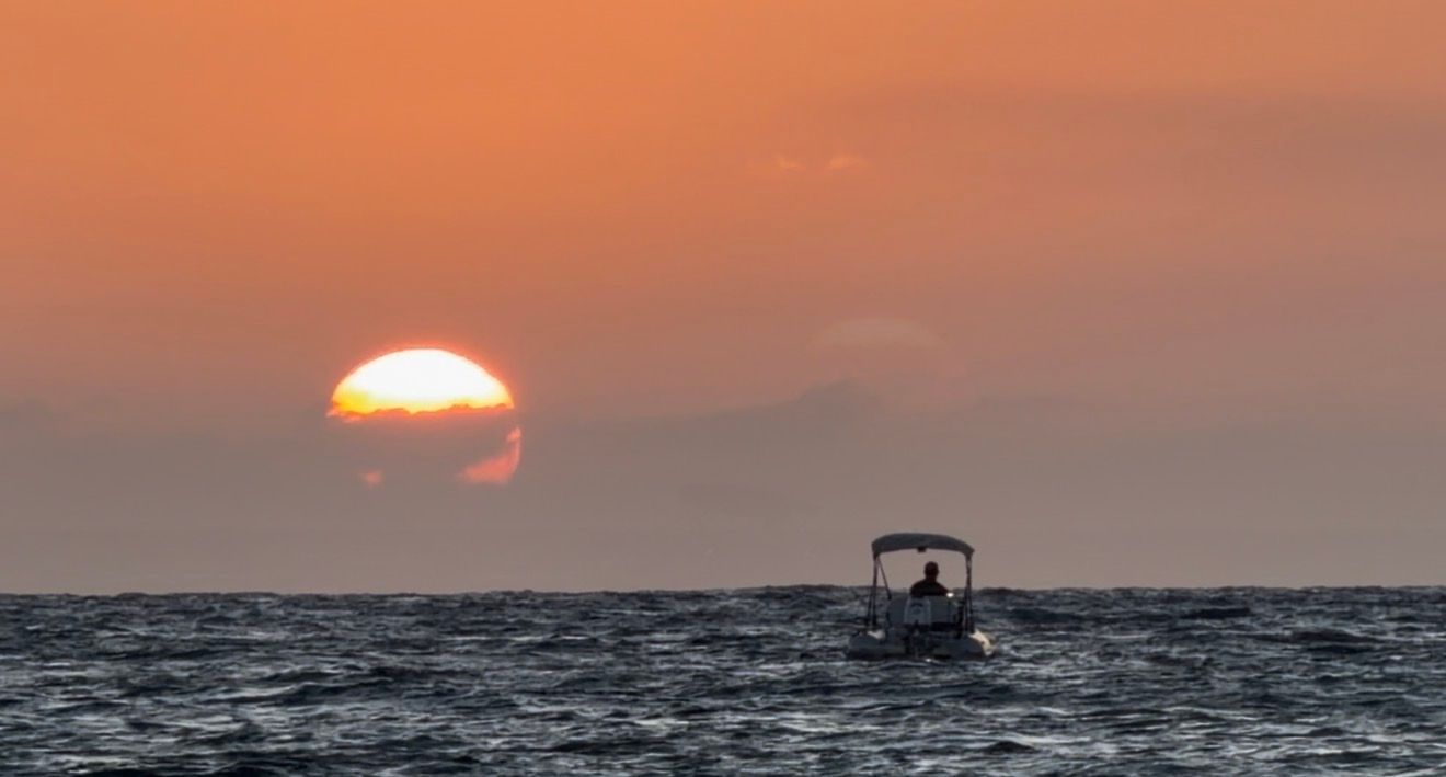 A small boat silhouetted against a vibrant orange sunset over calm ocean waters at dusk.