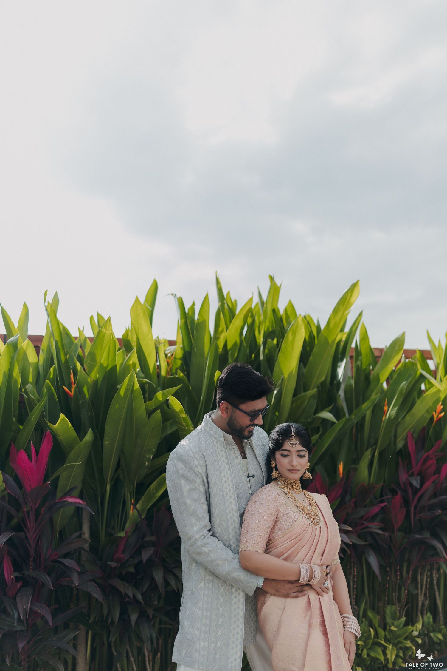 Indian couple in traditional wedding attire posing together in front of lush tropical greenery on their wedding day.