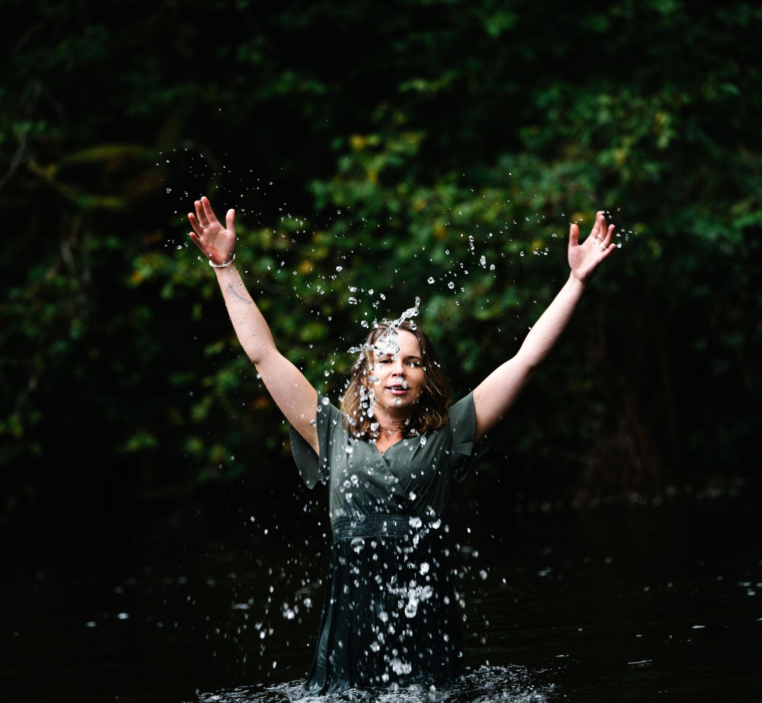 Woman standing in water with arms raised, surrounded by splashing water and lush green trees in background.