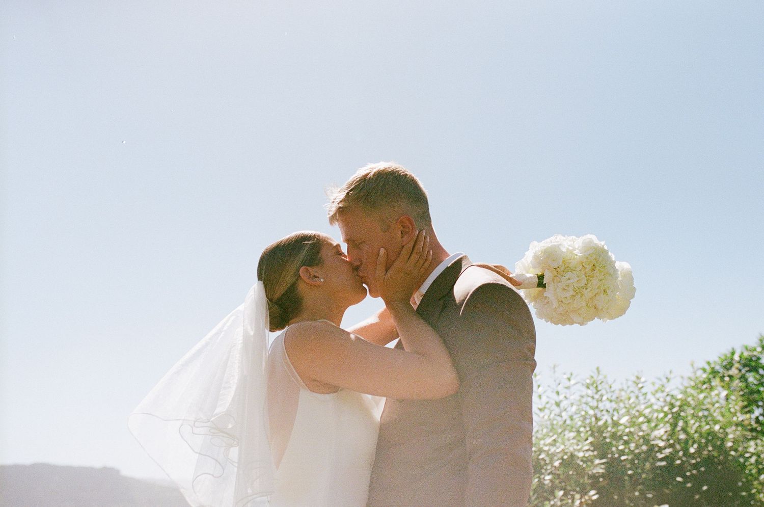 A couple embraces in a romantic outdoor setting with sunlight filtering through trees in the background.