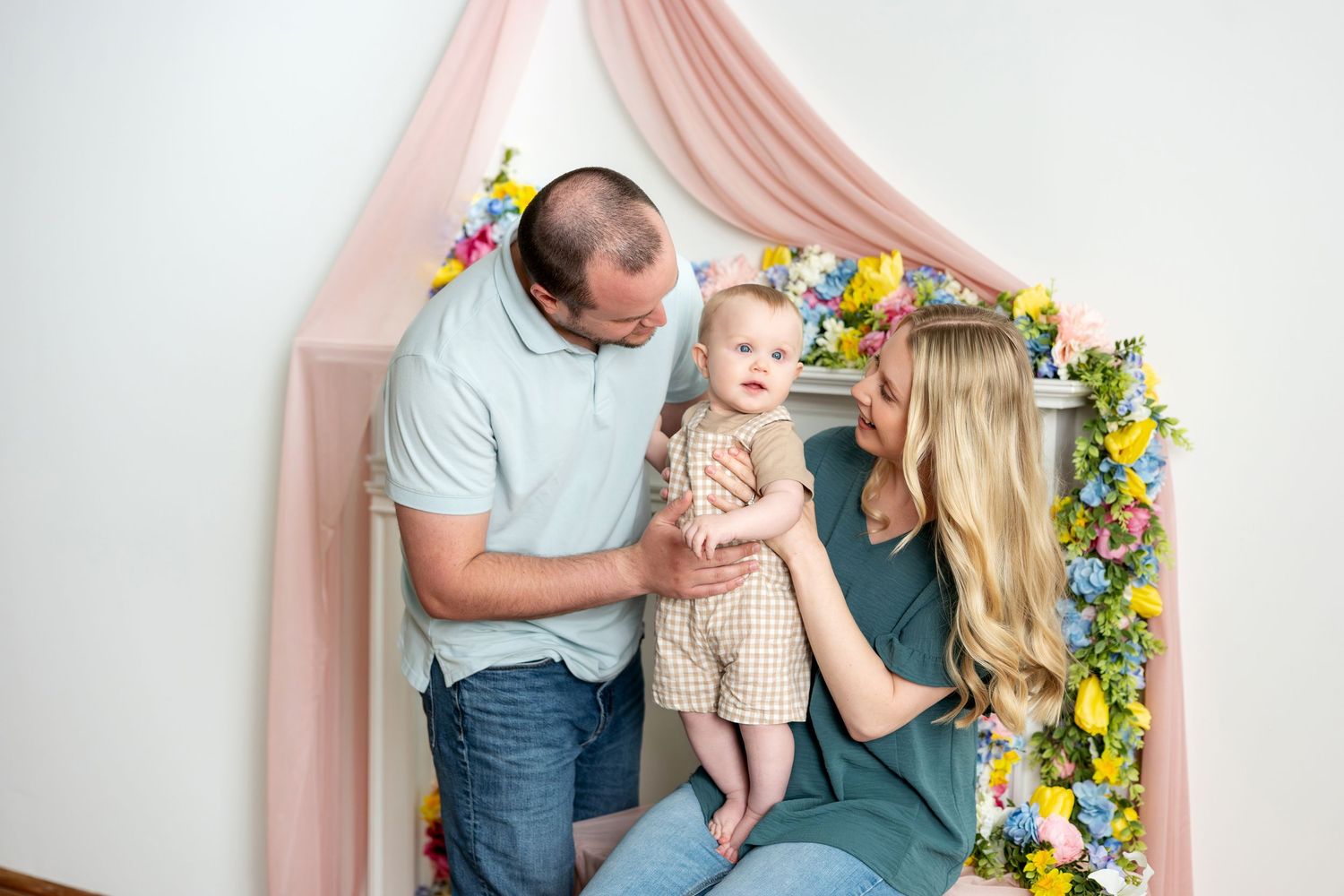 Happy family with baby in floral spring photo studio with pink drape backdrop.