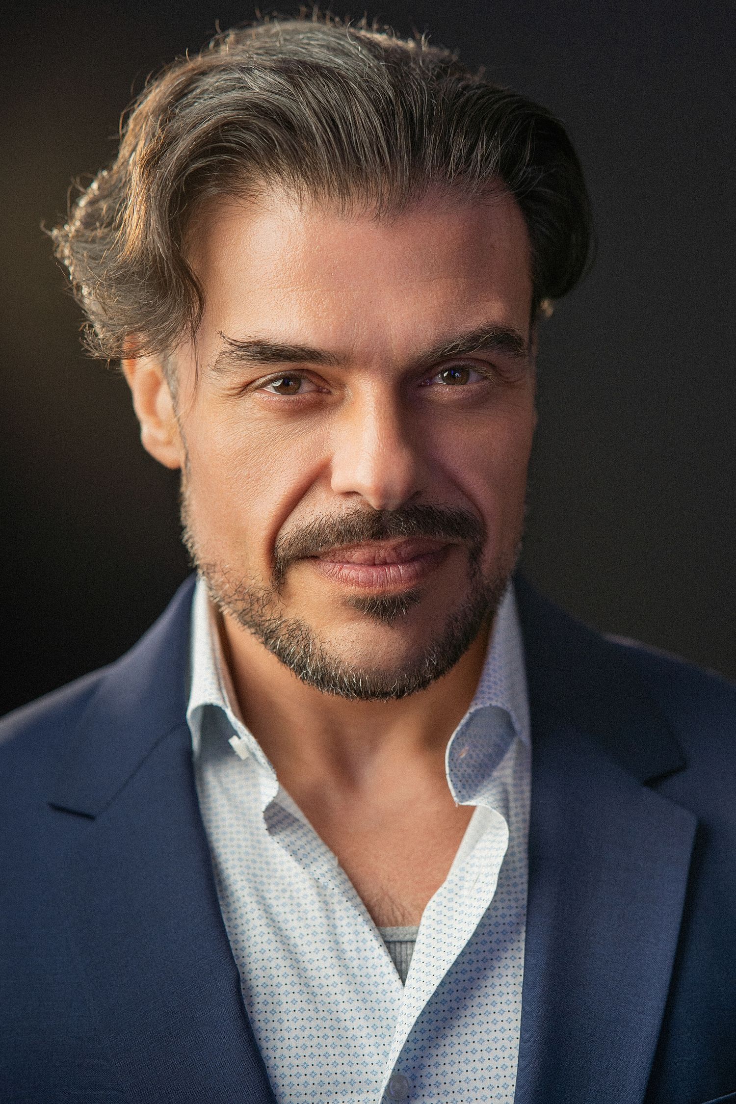 Professional headshot of a confident middle-aged man with dark hair, beard, wearing a blue suit jacket against dark background.
