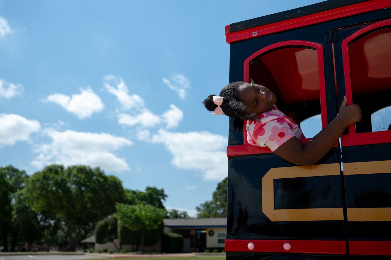 Young girl leaning out of a colorful red and black train ride on a sunny day with blue skies.