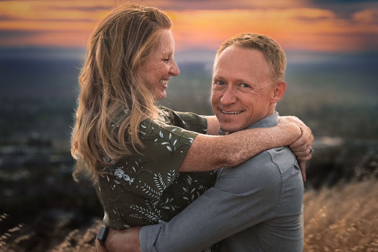 A smiling couple embraces at sunset with a scenic landscape backdrop, capturing a romantic outdoor moment.