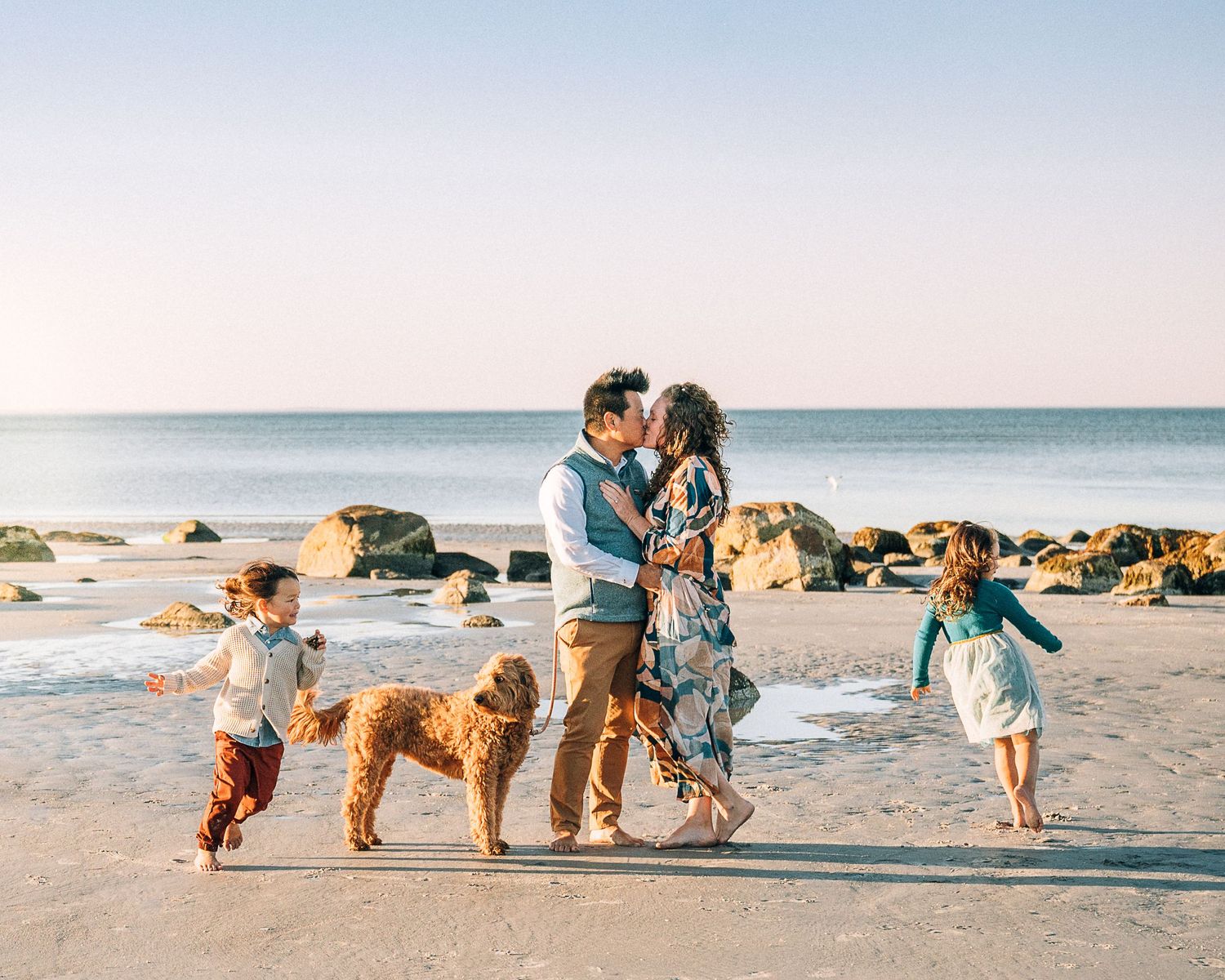Family with two kids and a golden doodle kissing on a sunny beach with rocks and calm ocean waves.