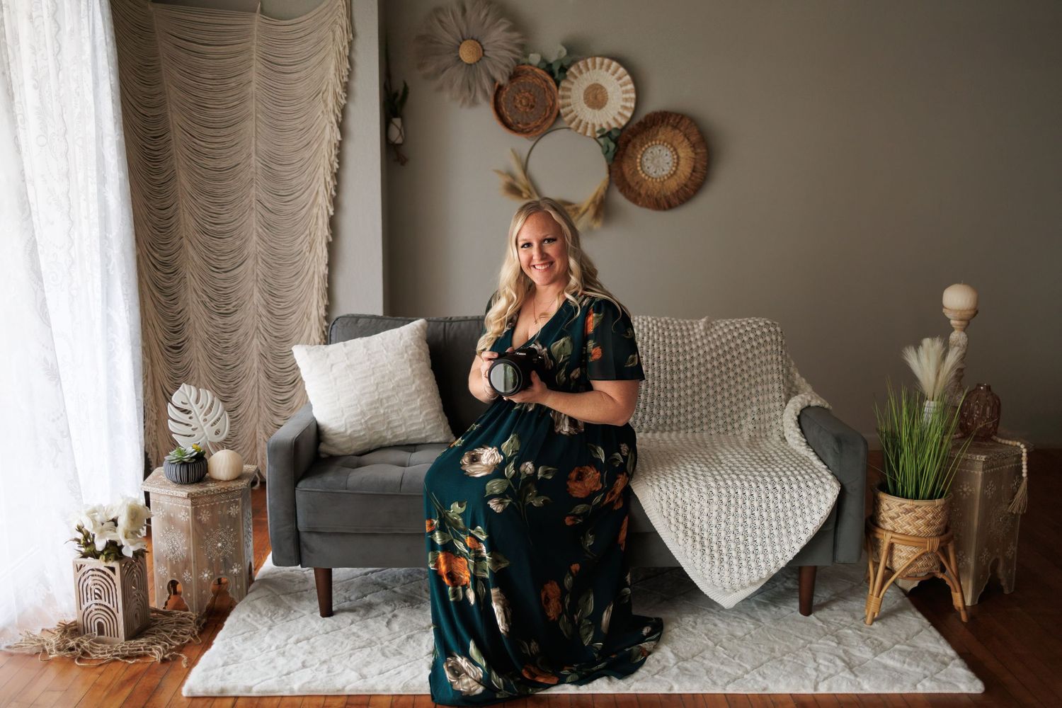 A self portrait of a photographer in a floral dress sitting on a gray couch in a modern living room with neutral decor and natural lighting.