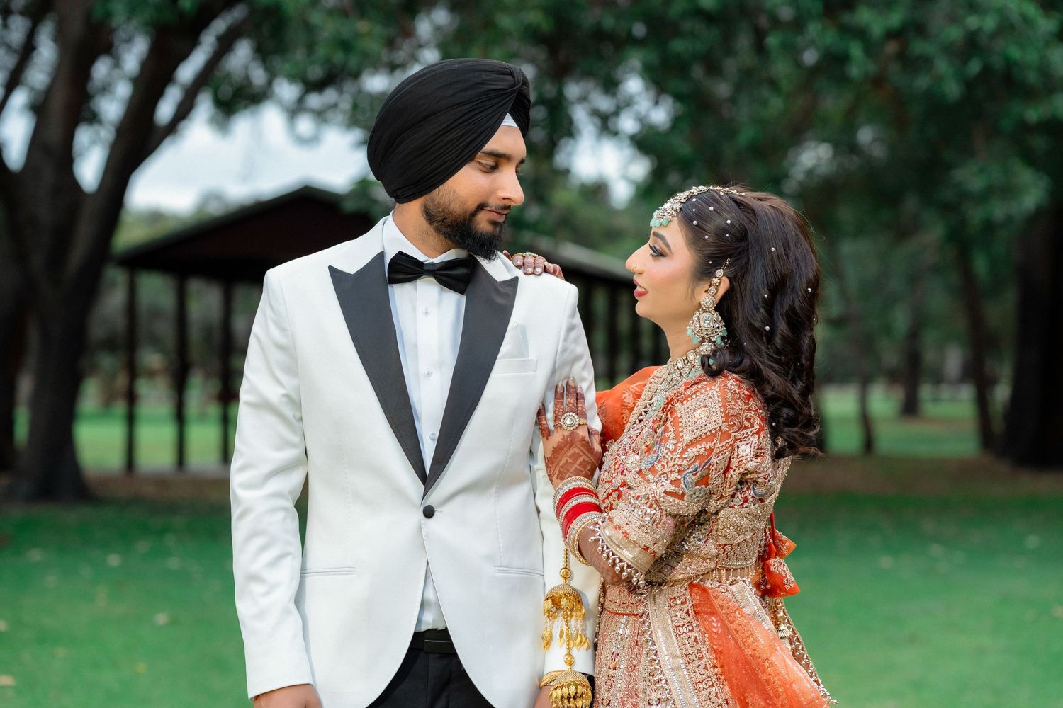 Sikh bride and groom in traditional wedding attire, sharing a romantic glance outdoors.