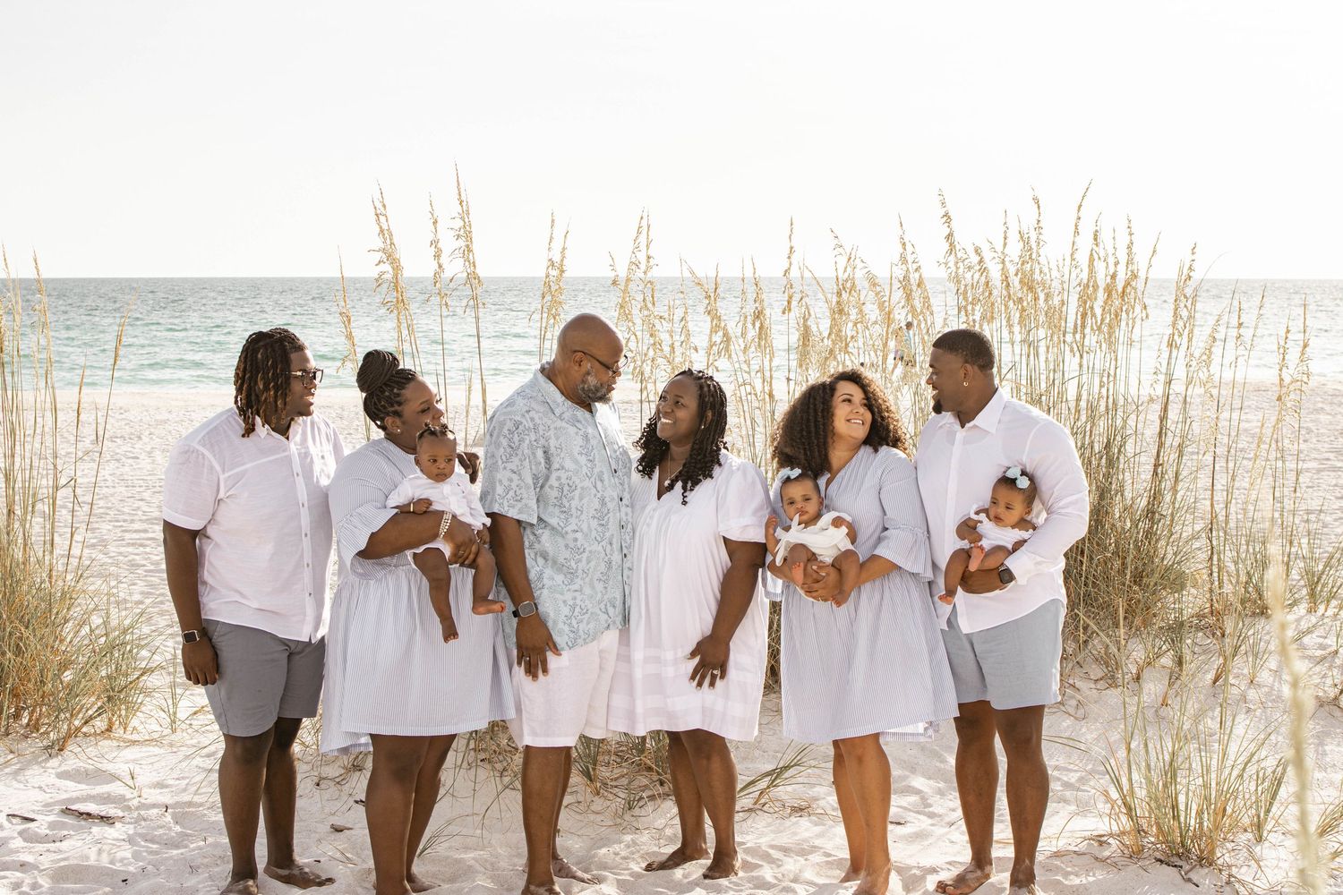 Family portrait of seven people in white outfits on a sandy beach with sea oats and ocean in background.