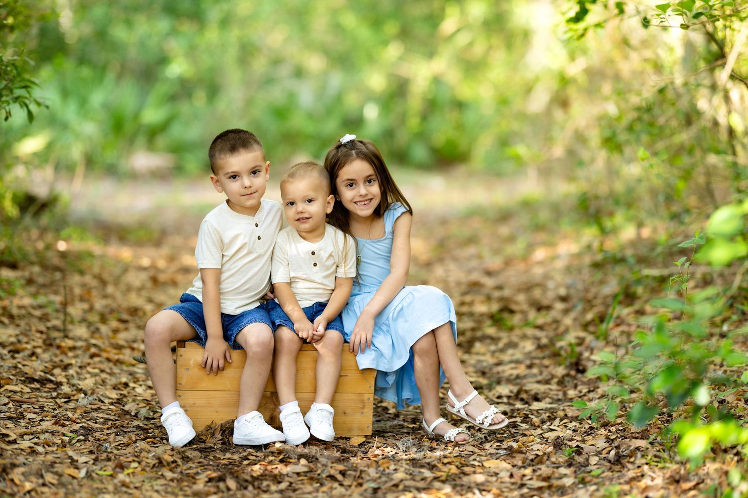 Three young children sitting together on a wooden crate on a forest path surrounded by green trees.