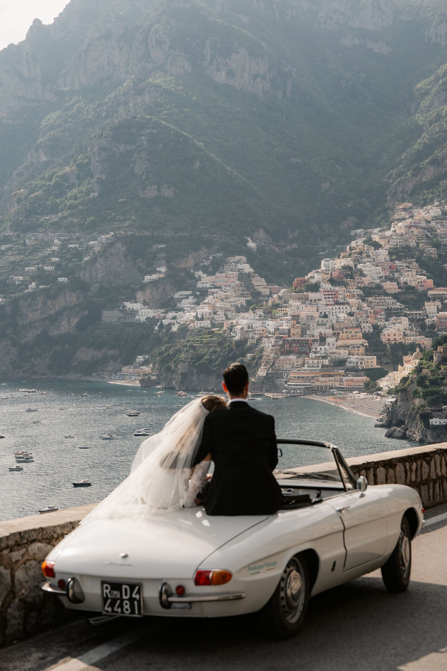 Bride and groom sit on a vintage white convertible overlooking Positano, Italy's cliffside village and coastline.