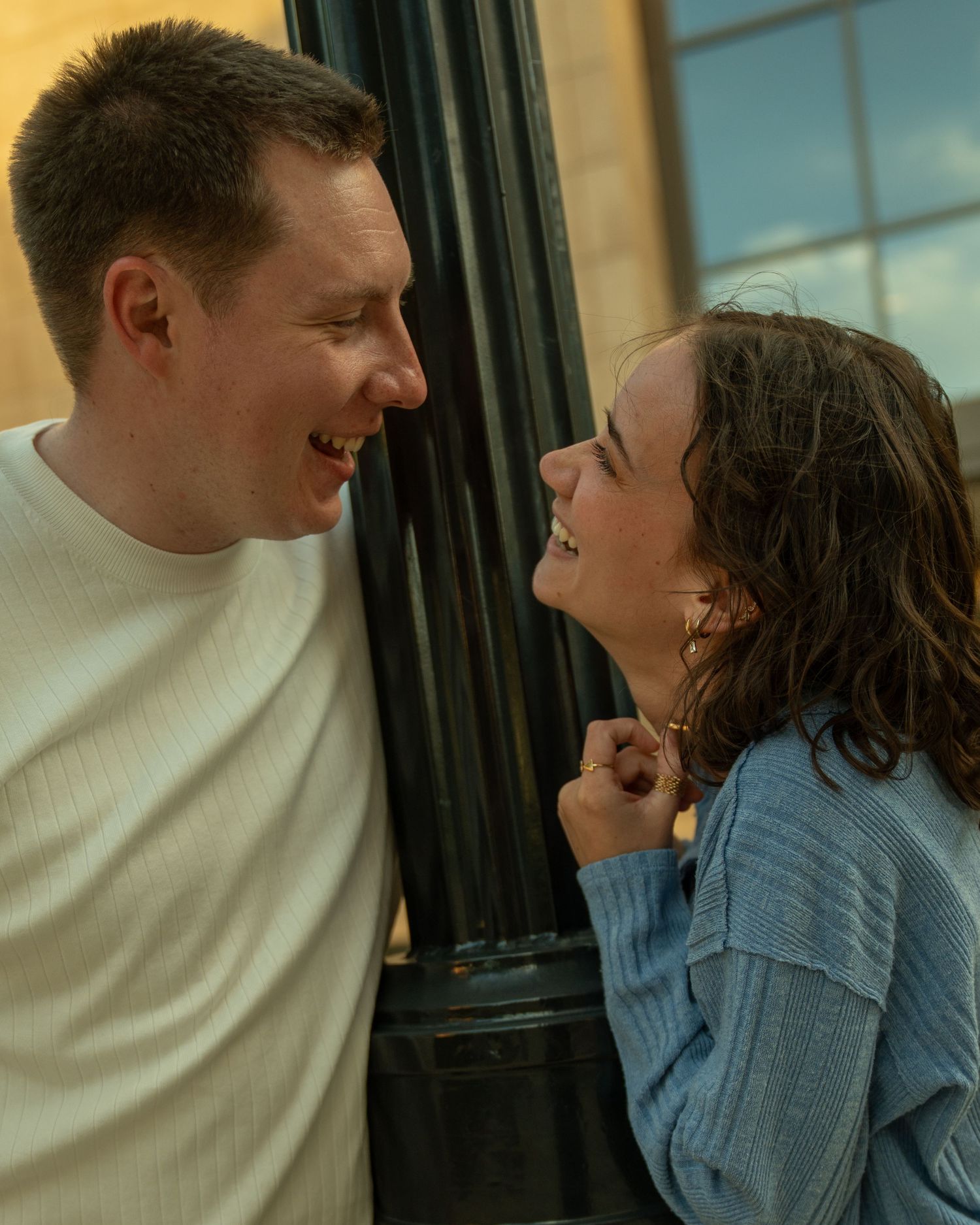 A smiling couple sharing a joyful moment, leaning close together outdoors near a black pillar.