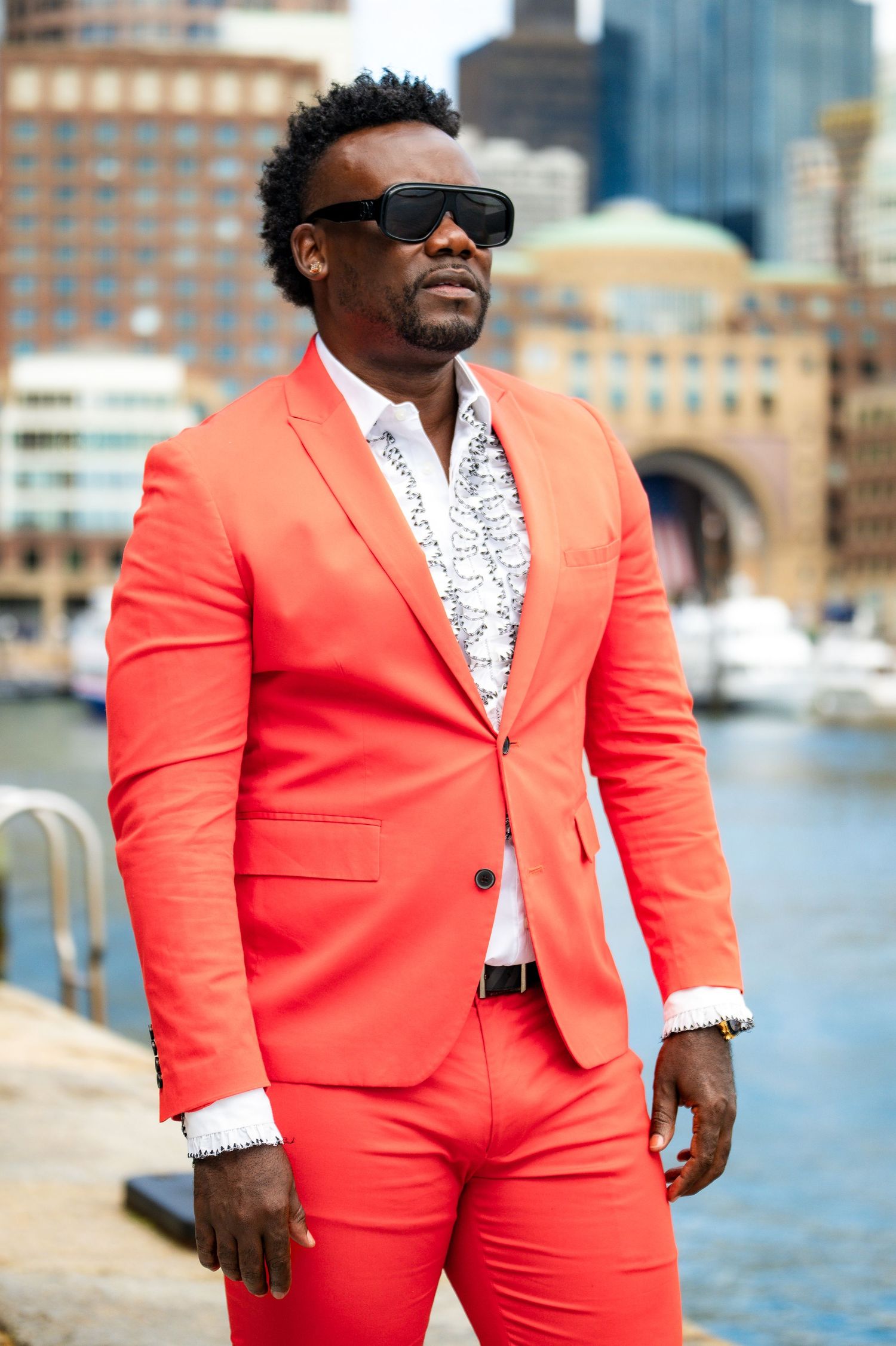 Man in bold coral red suit and sunglasses posing outdoors near waterfront with city buildings in background.