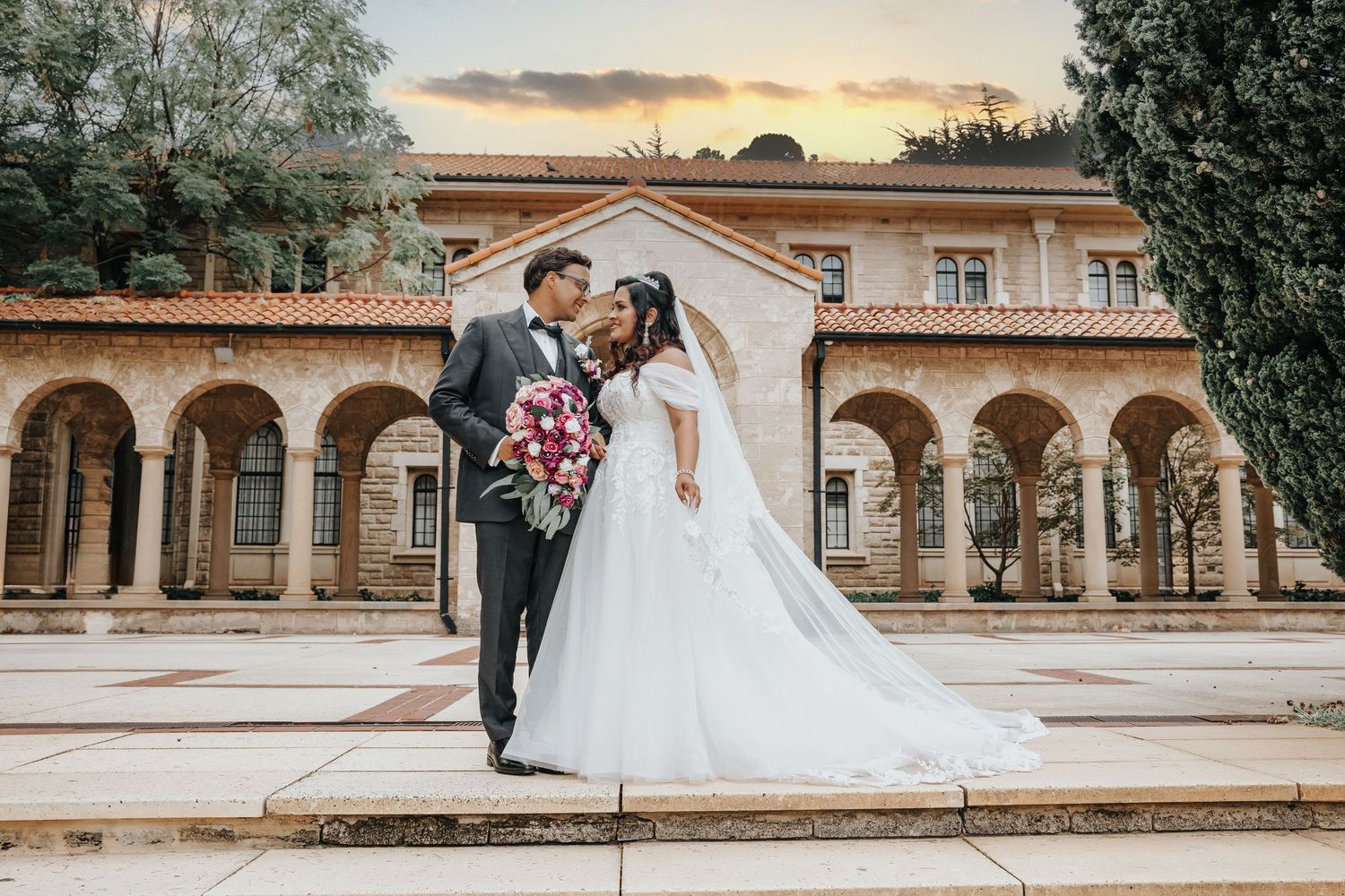 Bride and groom share a romantic moment in front of a historic arched building at sunset.
