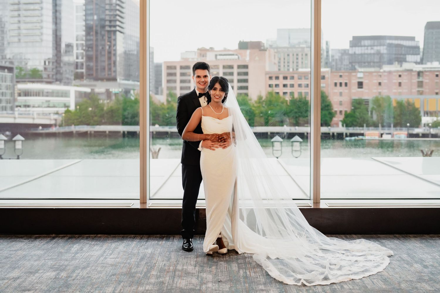 Bride in white gown with cathedral veil stands with groom in black tuxedo before large city-view windows.
