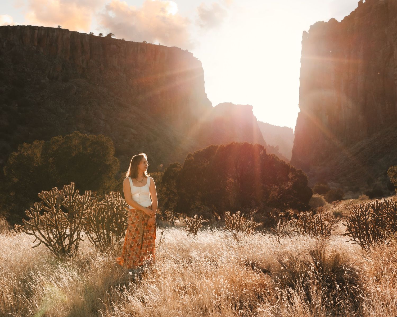 Young woman in white lace dress poses in desert canyon with golden sunset lens flare behind her.