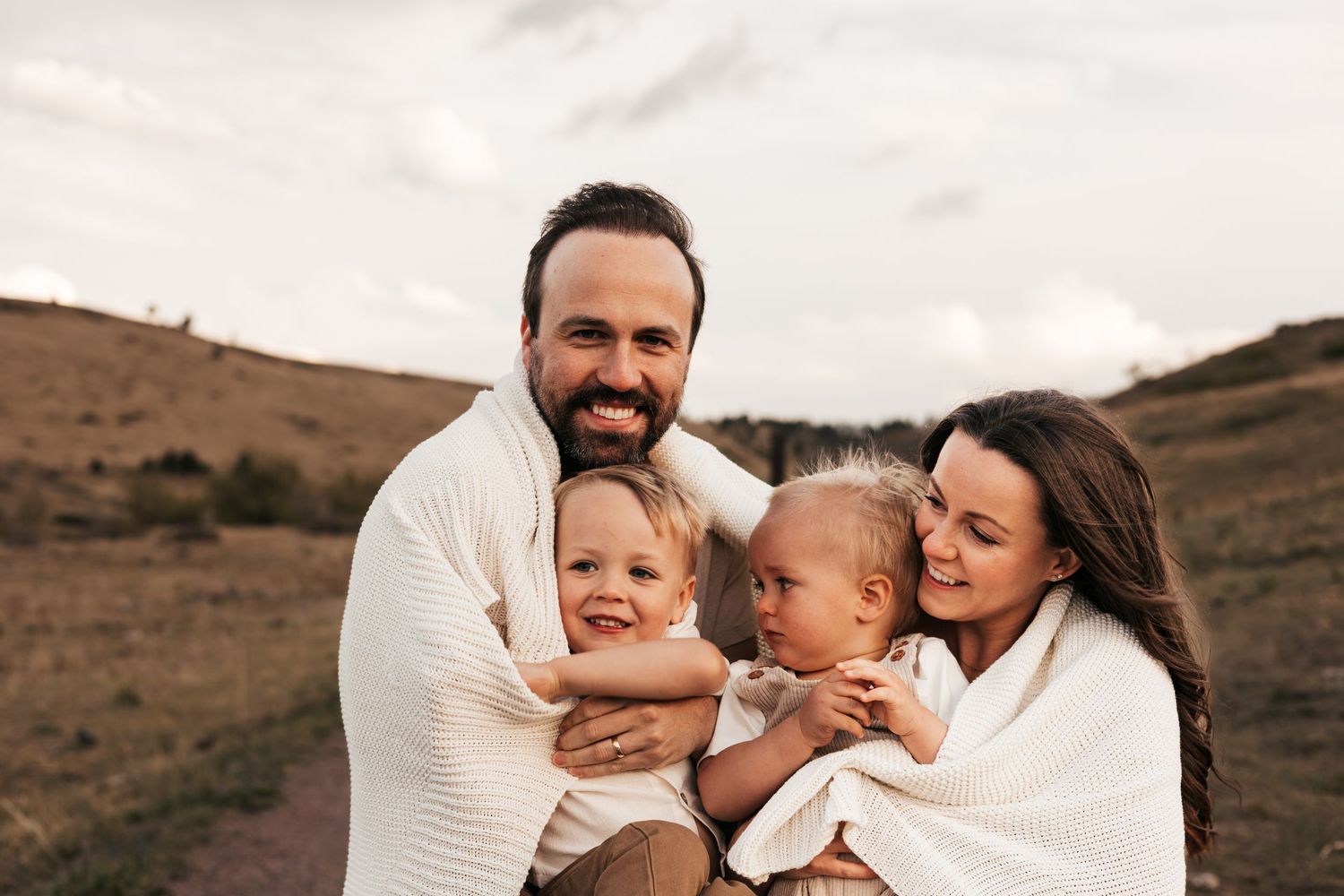 Happy family of four in white sweaters posing outdoors in a field, parents holding two young toddlers.