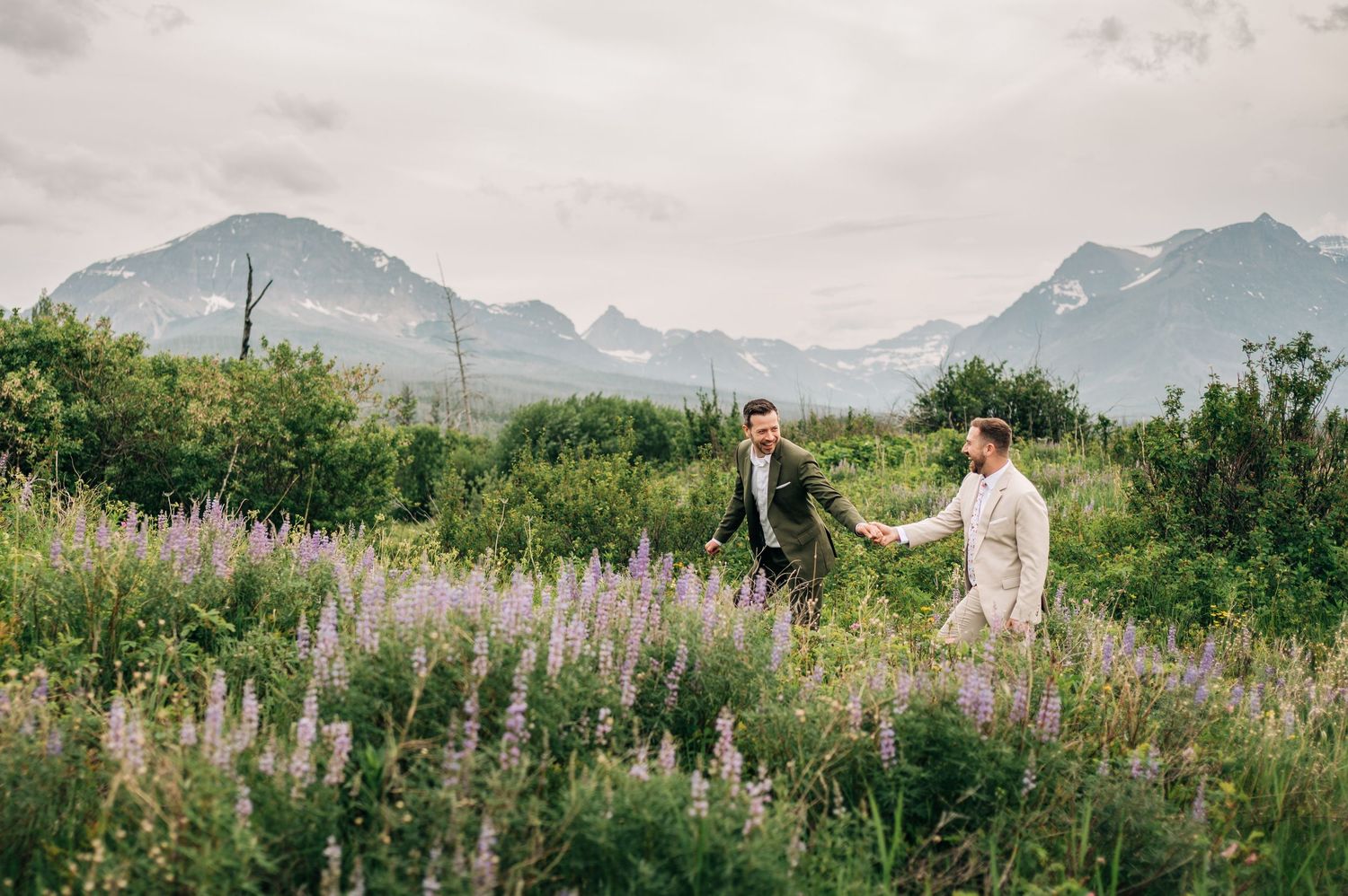 Two grooms hold hands in a wildflower meadow with majestic mountain peaks in the background.