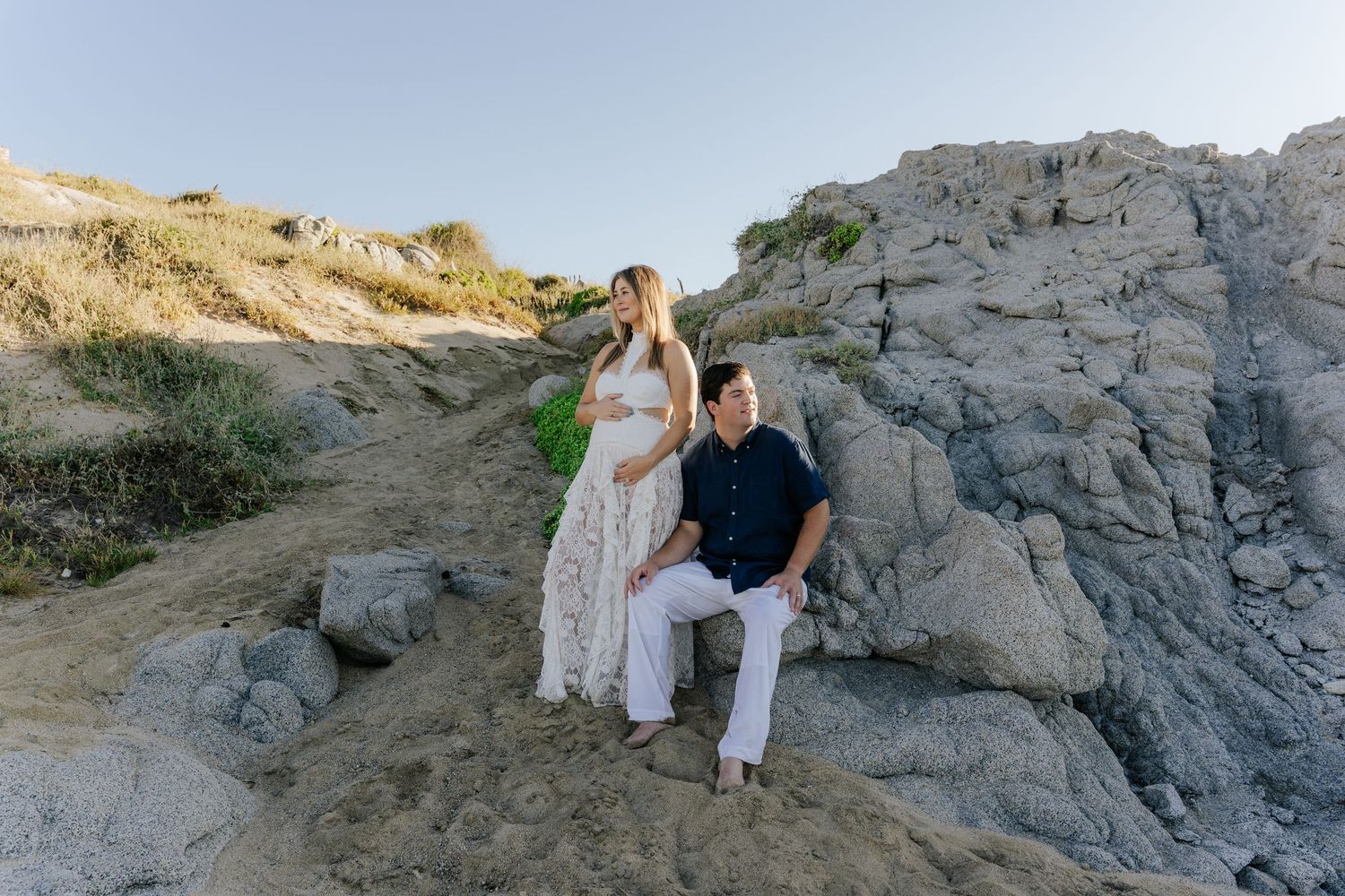 Pregnant woman in floral dress with partner sitting on coastal rocks during golden hour maternity photo session.