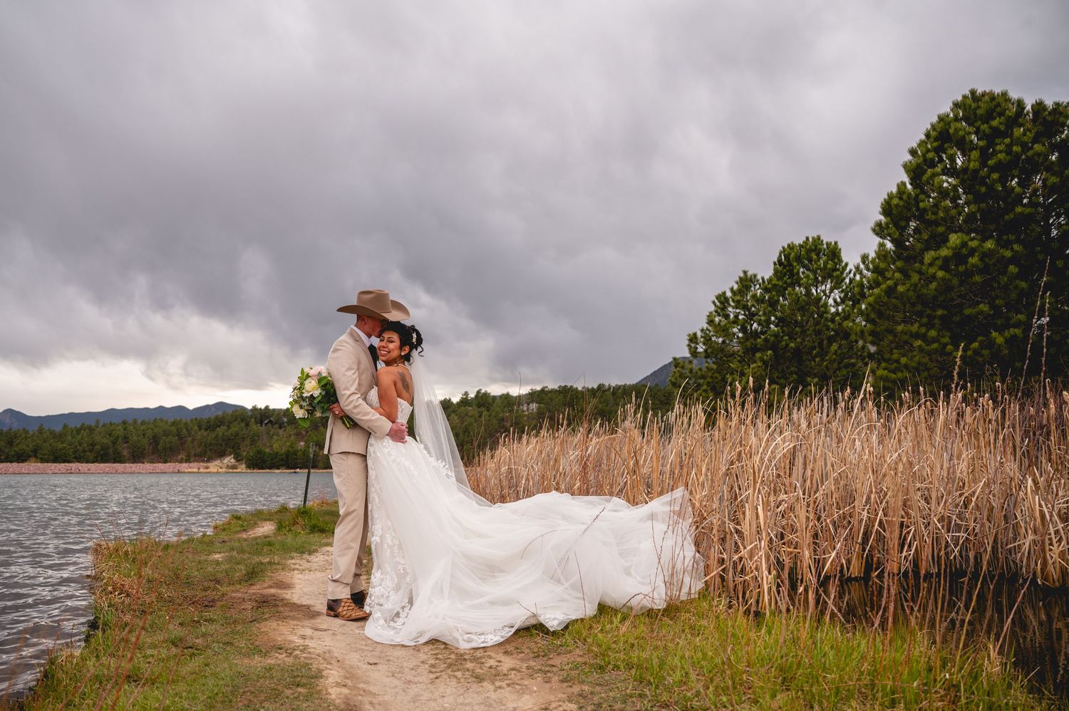 Bride and groom share a romantic kiss by a lakeside with dramatic stormy skies and tall reeds in background.