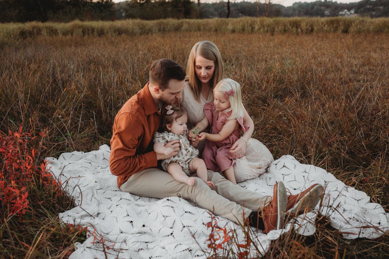 A couple sits together on a blanket outdoors during golden hour with their white dog in autumn.