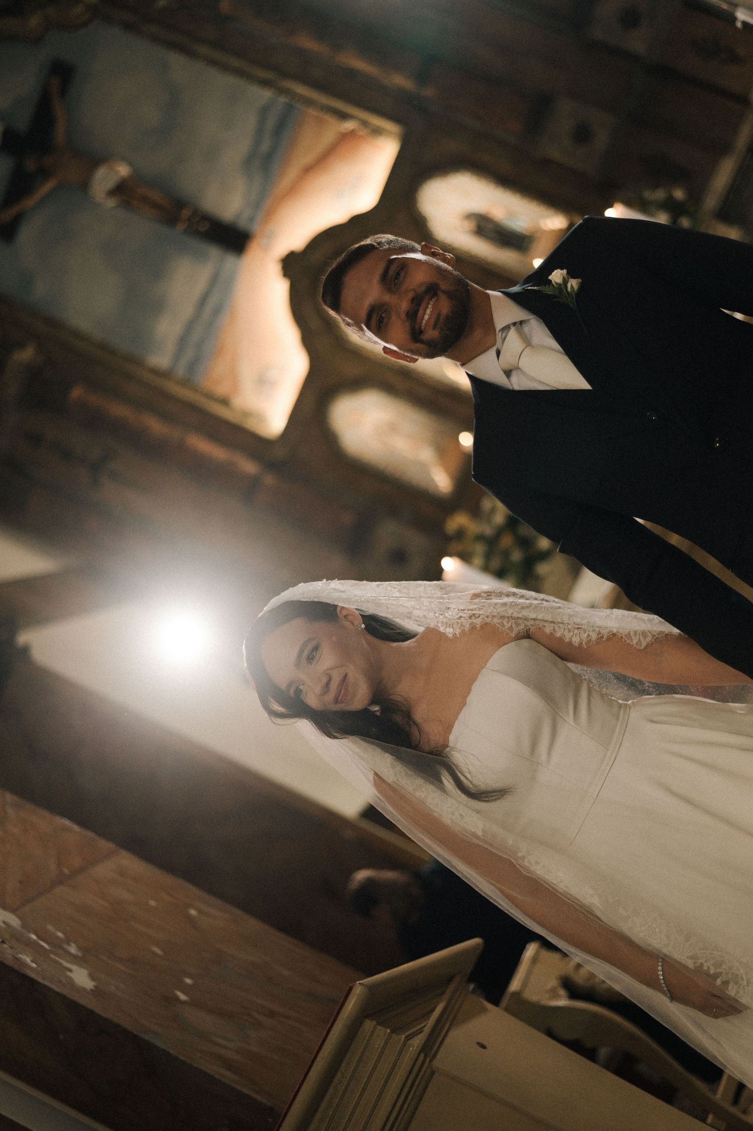 A smiling bride in a white gown and veil walks with her groom in a black suit inside a historic church.