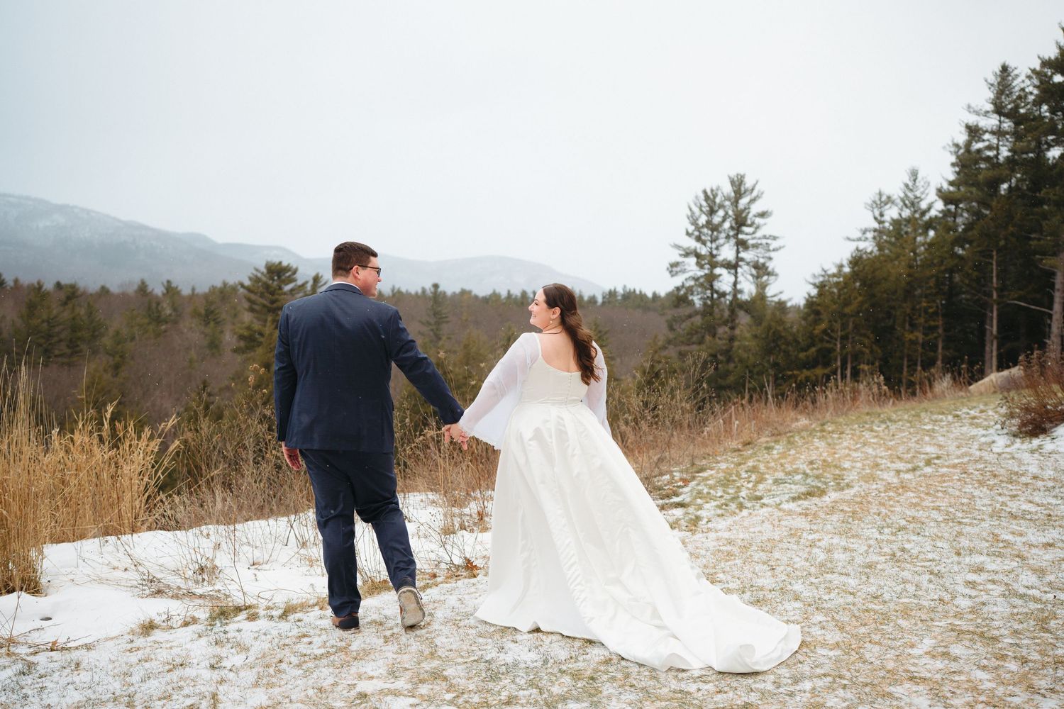 Couple in wedding attire walking through a snowy mountain landscape during their winter wedding photoshoot.