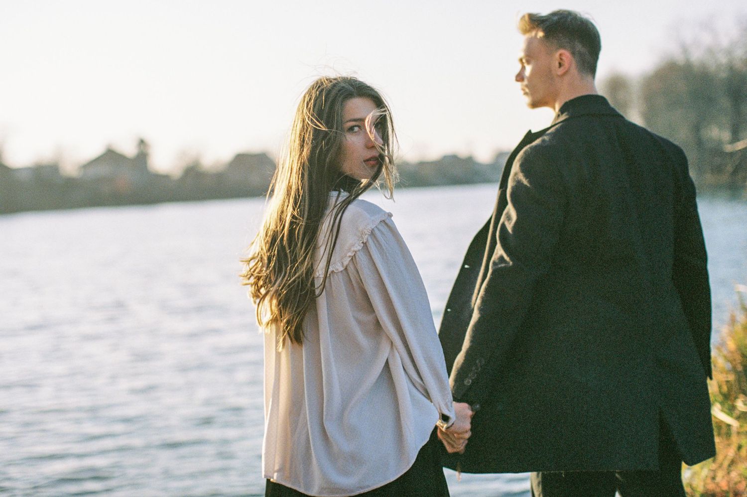 A couple stands together holding hands by a serene lake at sunset with golden light illuminating the scene.
