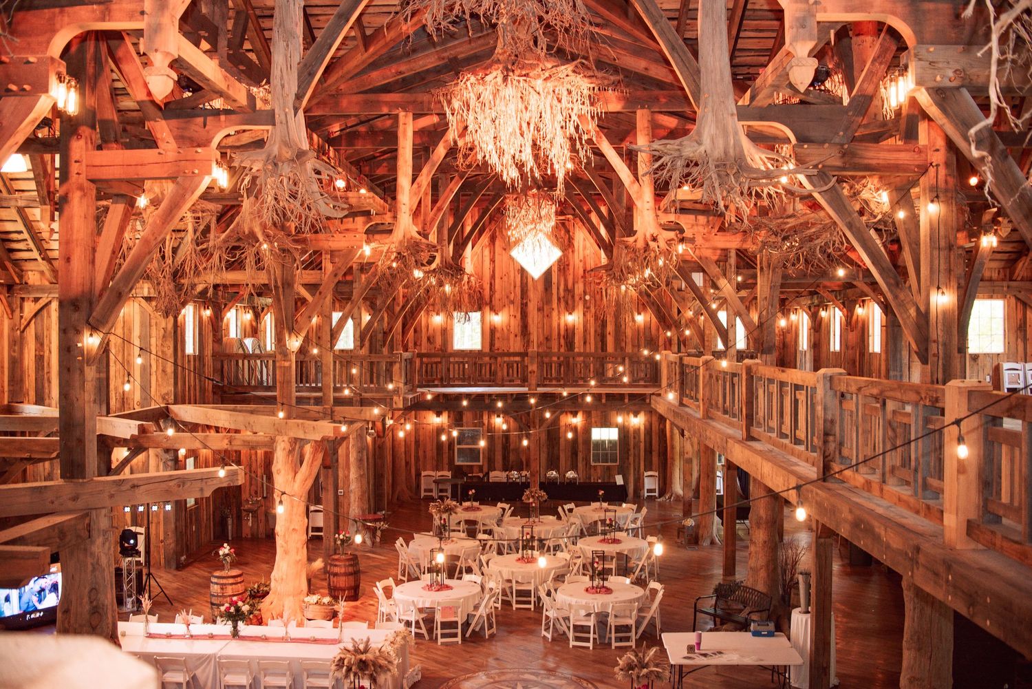 Rustic barn wedding venue with wooden beams, antler chandeliers, string lights, and elegantly set round tables for a reception.