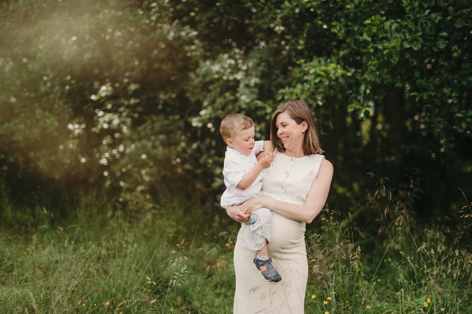 A mother in a white dress holds her baby outdoors in a sunlit green garden setting.