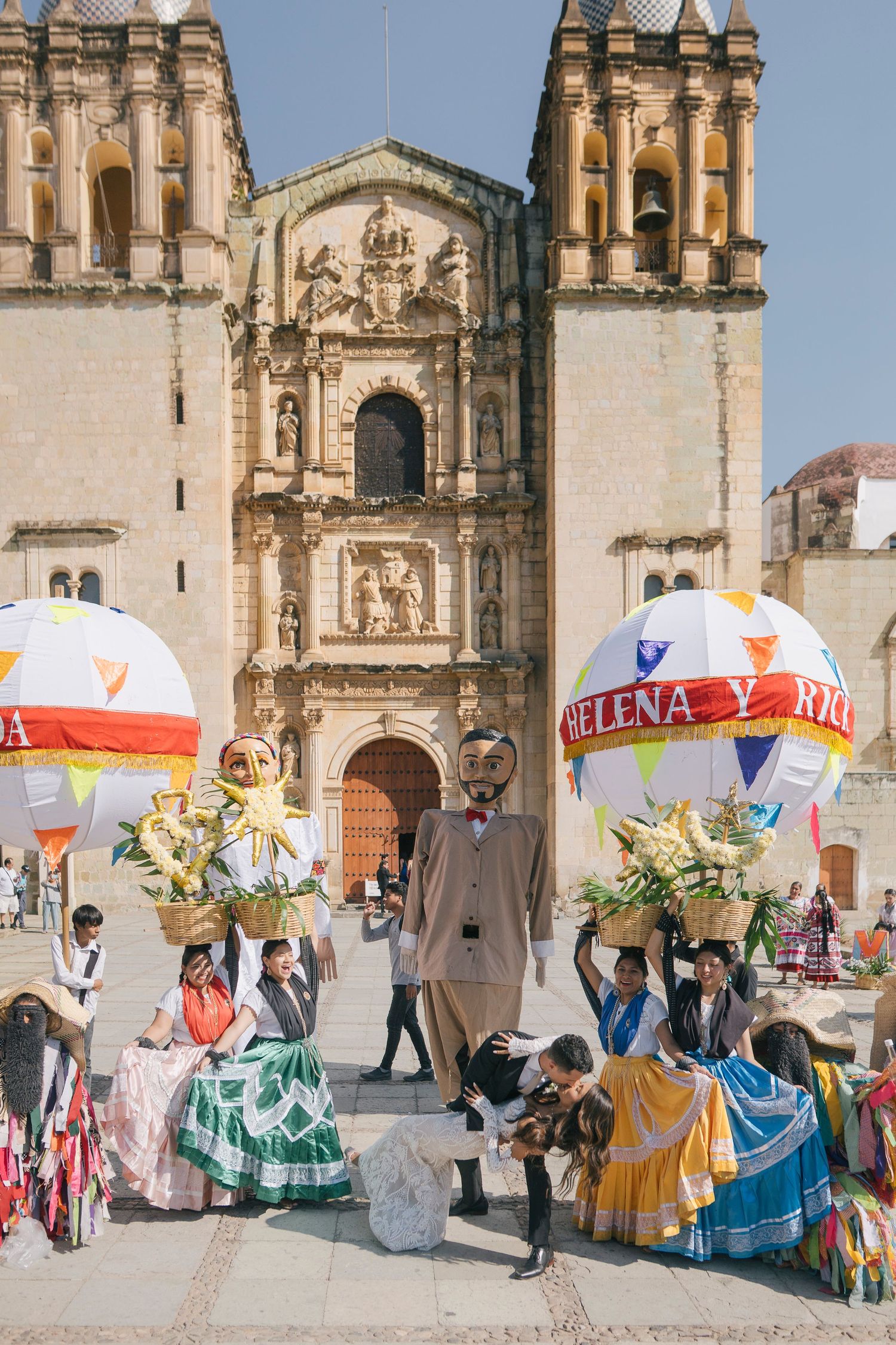 Colorful Mexican festival with giant puppets and dancers in traditional dress outside a historic baroque church in Oaxaca.