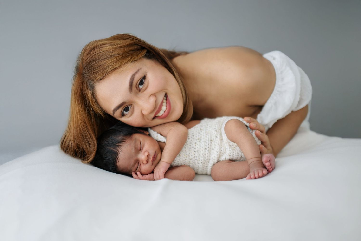 Smiling mother lying on white bed posing with her sleeping newborn baby wrapped in a knit white outfit.