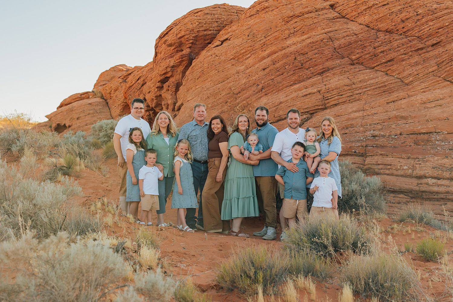 Large family posing together in front of red rock formations in a desert landscape, wearing coordinated teal and white outfits.