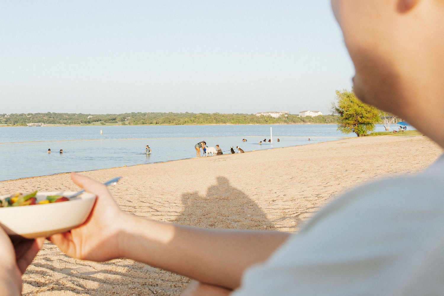 Person holding a bowl of food on a sunny sandy beach with calm water and people relaxing in the background.