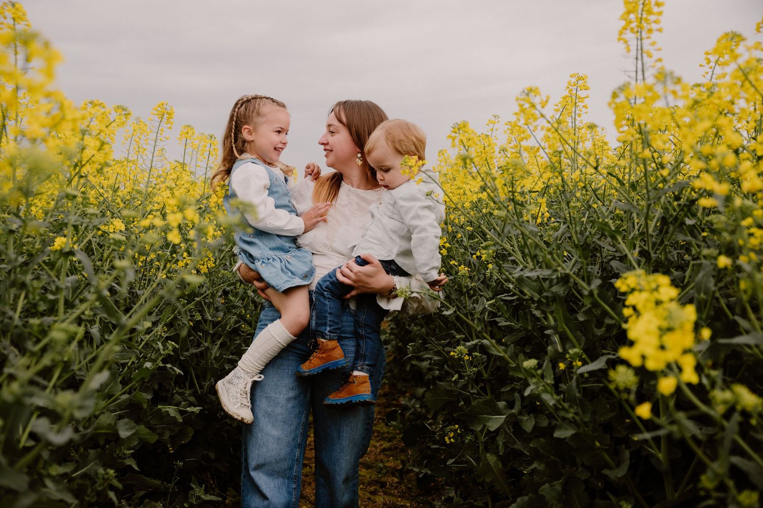 Mother holding two young children in a vibrant yellow rapeseed flower field on an overcast day.