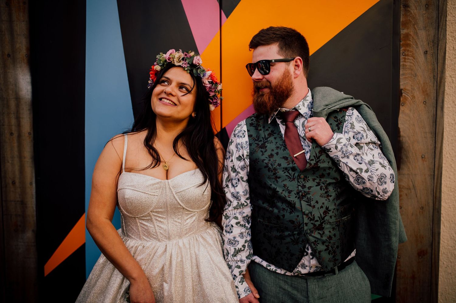 A couple in wedding attire laughs together against a colorful geometric wall backdrop at their celebration.