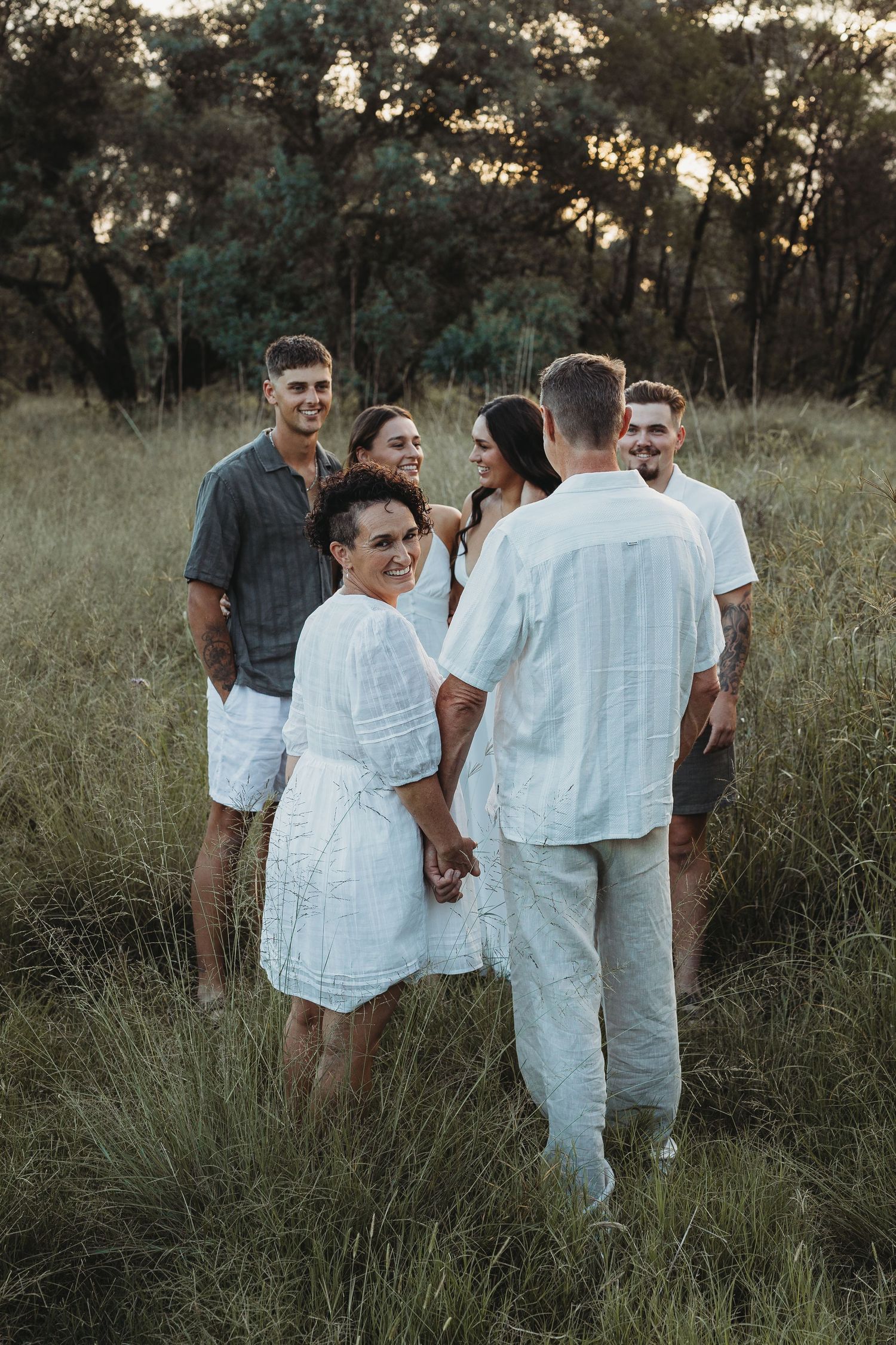 A group of people in light colored clothing gather together in a grassy field at sunset.