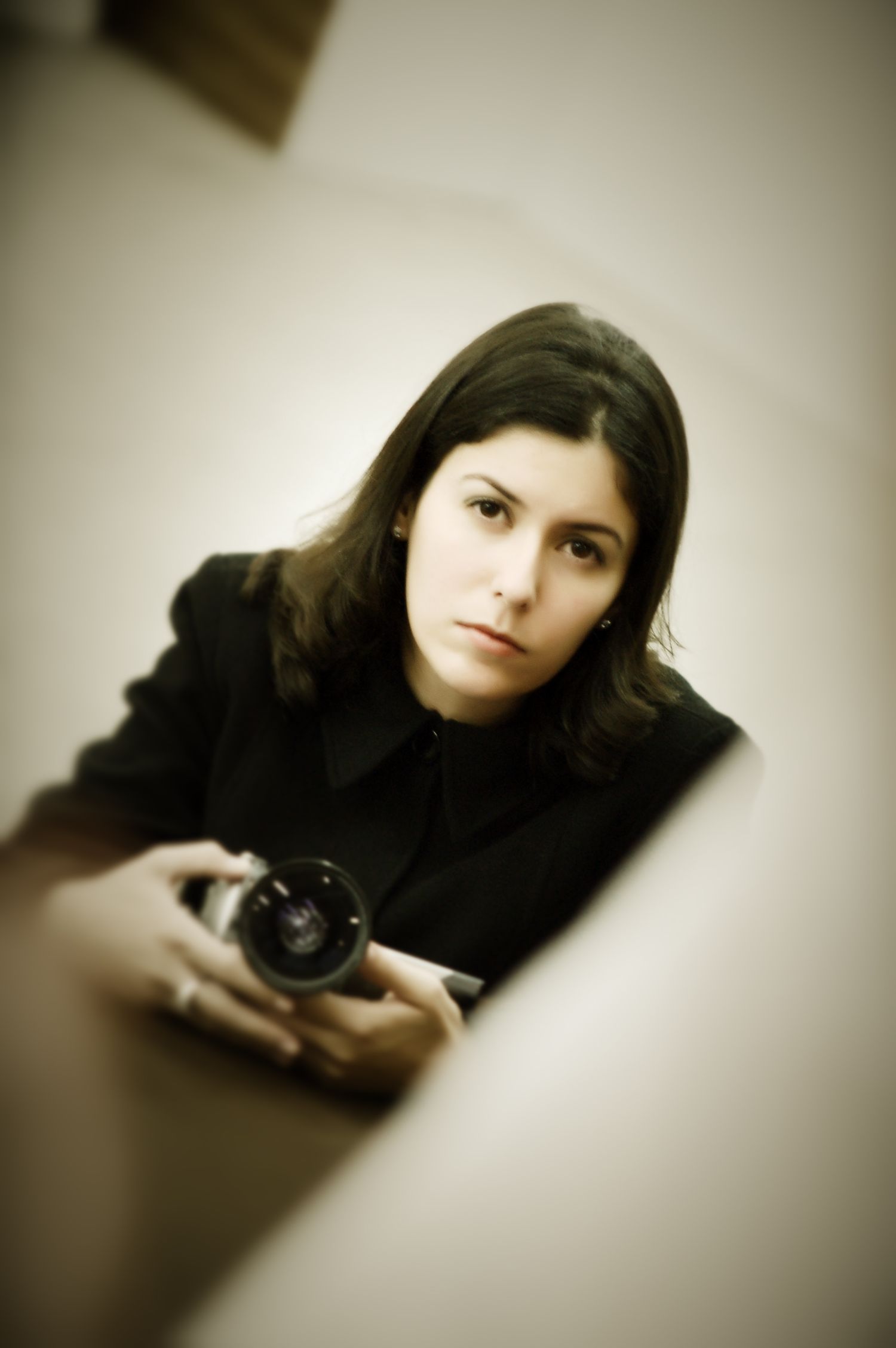Young woman in black jacket holding a camera, looking up with a serious expression in a softly lit room.