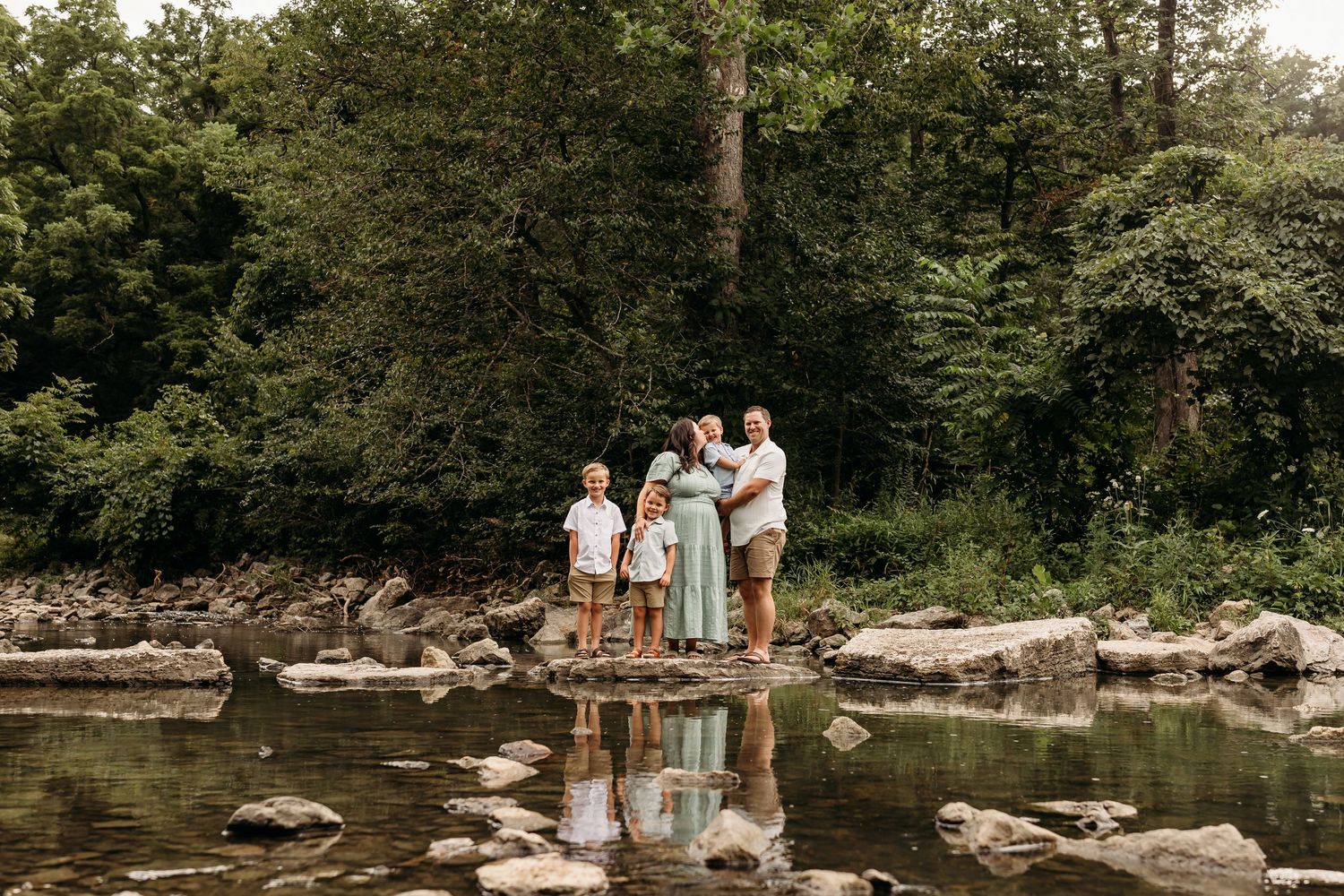 Family of four standing on rocks by a peaceful creek surrounded by lush green trees in a nature setting.