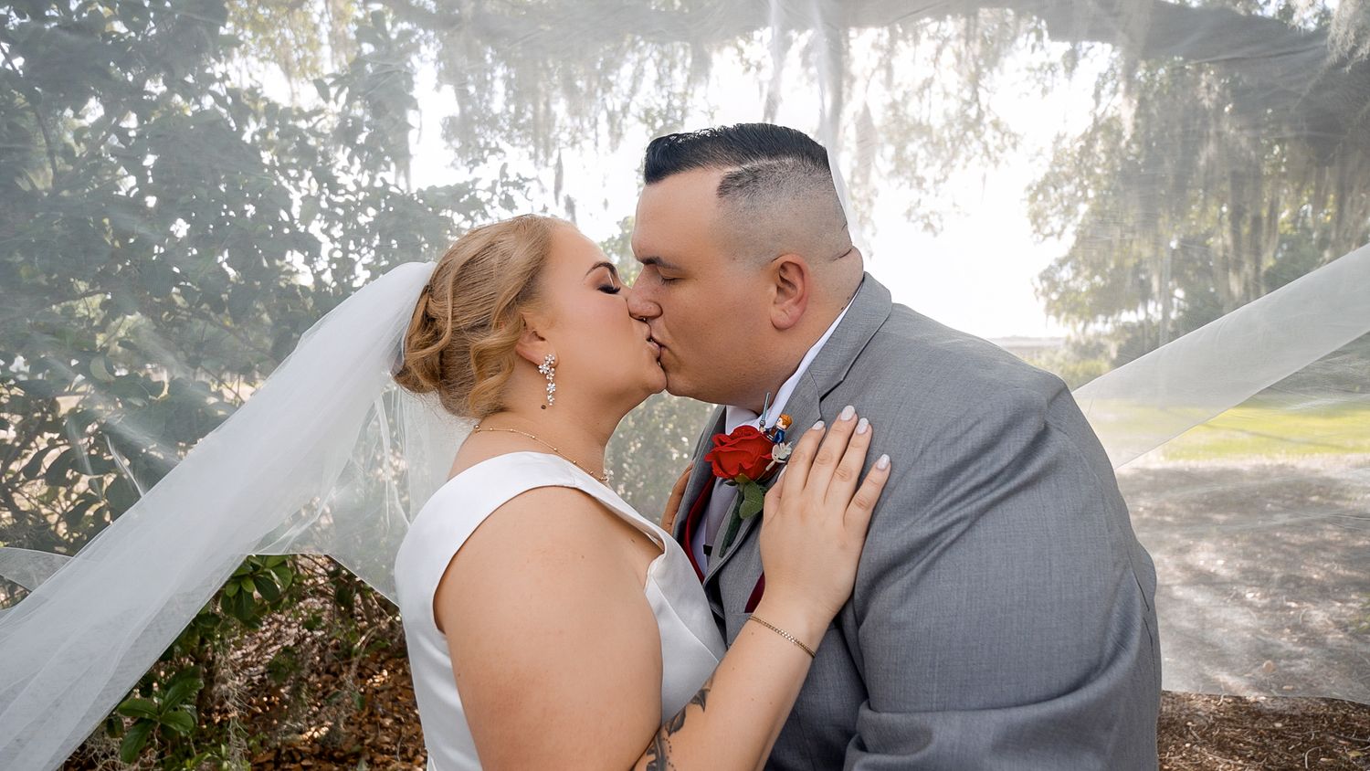 Bride and groom share a romantic kiss under a flowing veil outdoors, surrounded by moss-draped trees on their wedding day.