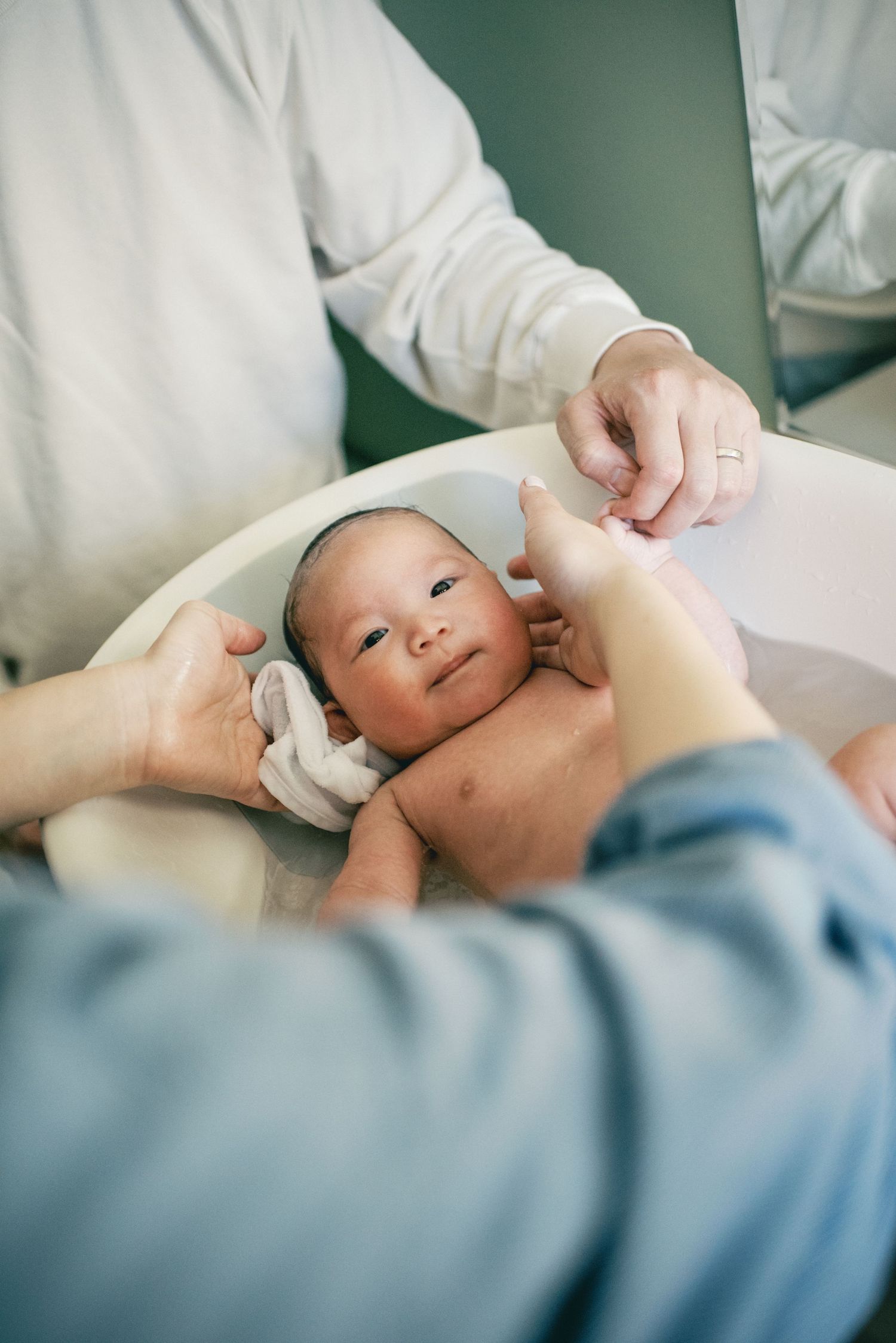 Newborn baby crying during first bath in white infant tub, supported by caregiver's hands.