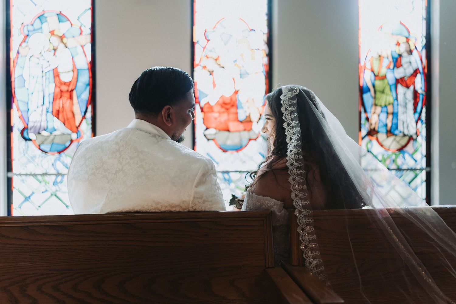 A couple sits together in a church pew with colorful stained glass windows in the background.