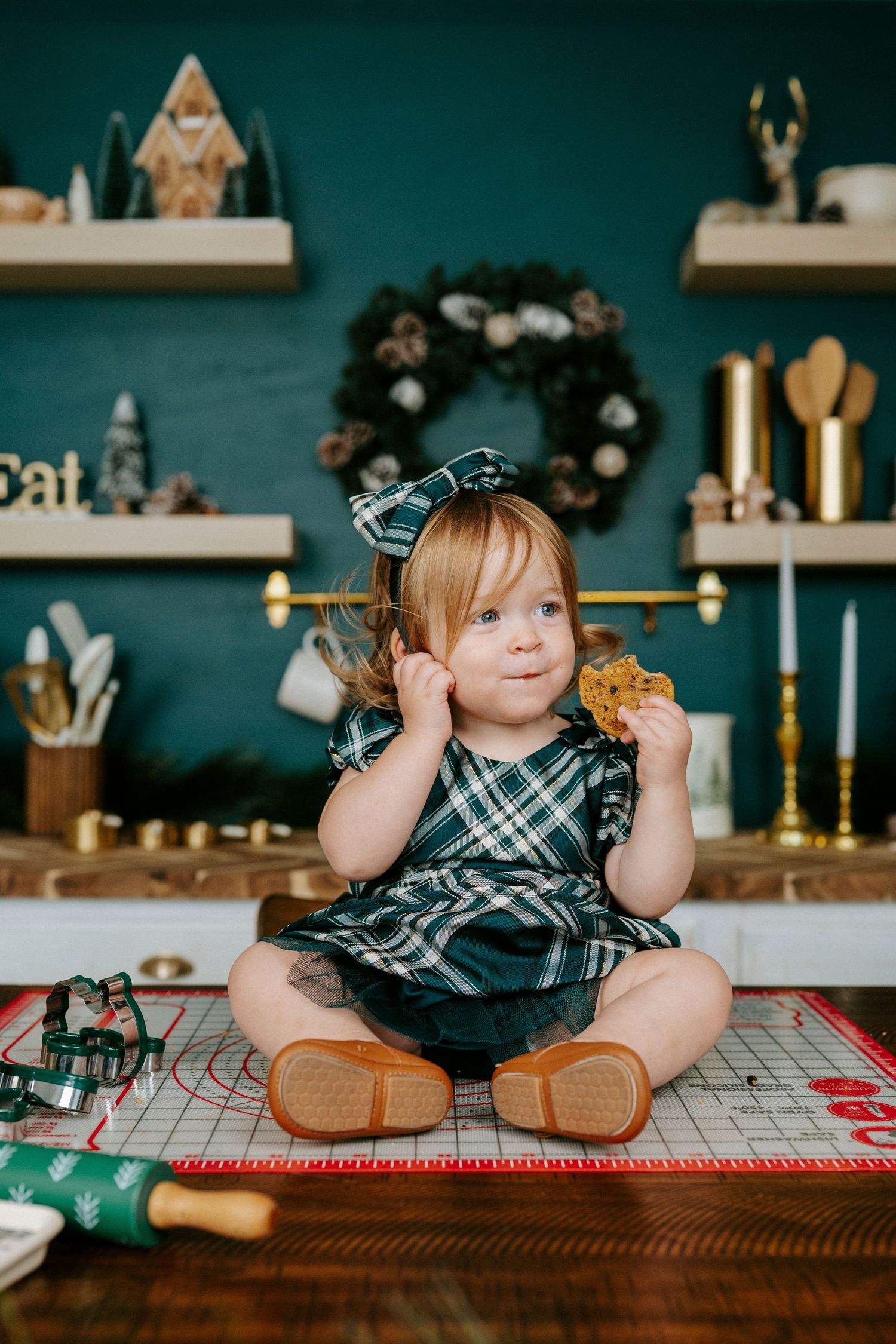 A child in a plaid dress sits on a wooden table with holiday decor and teal walls in the background.